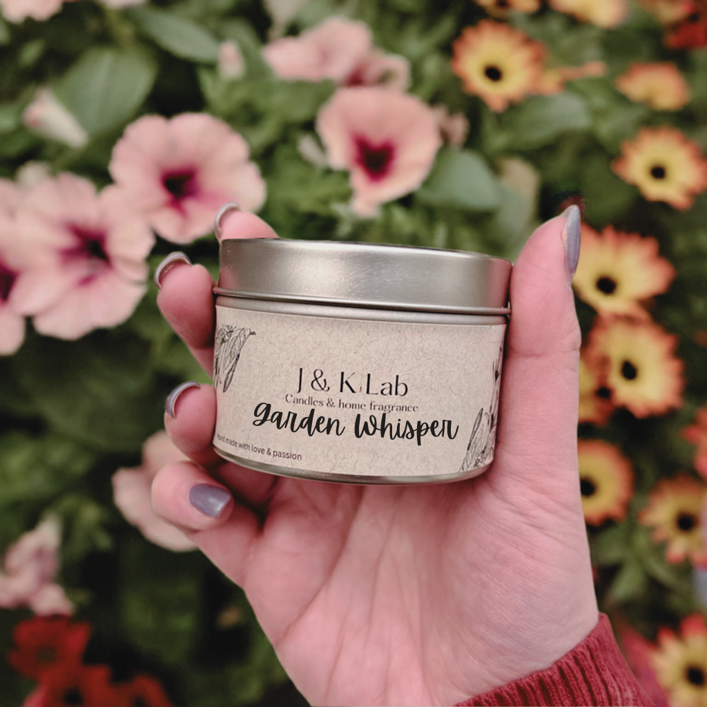 Hand holding a J&K Lab tin candle labeled 'Garden Whisper' against a floral background