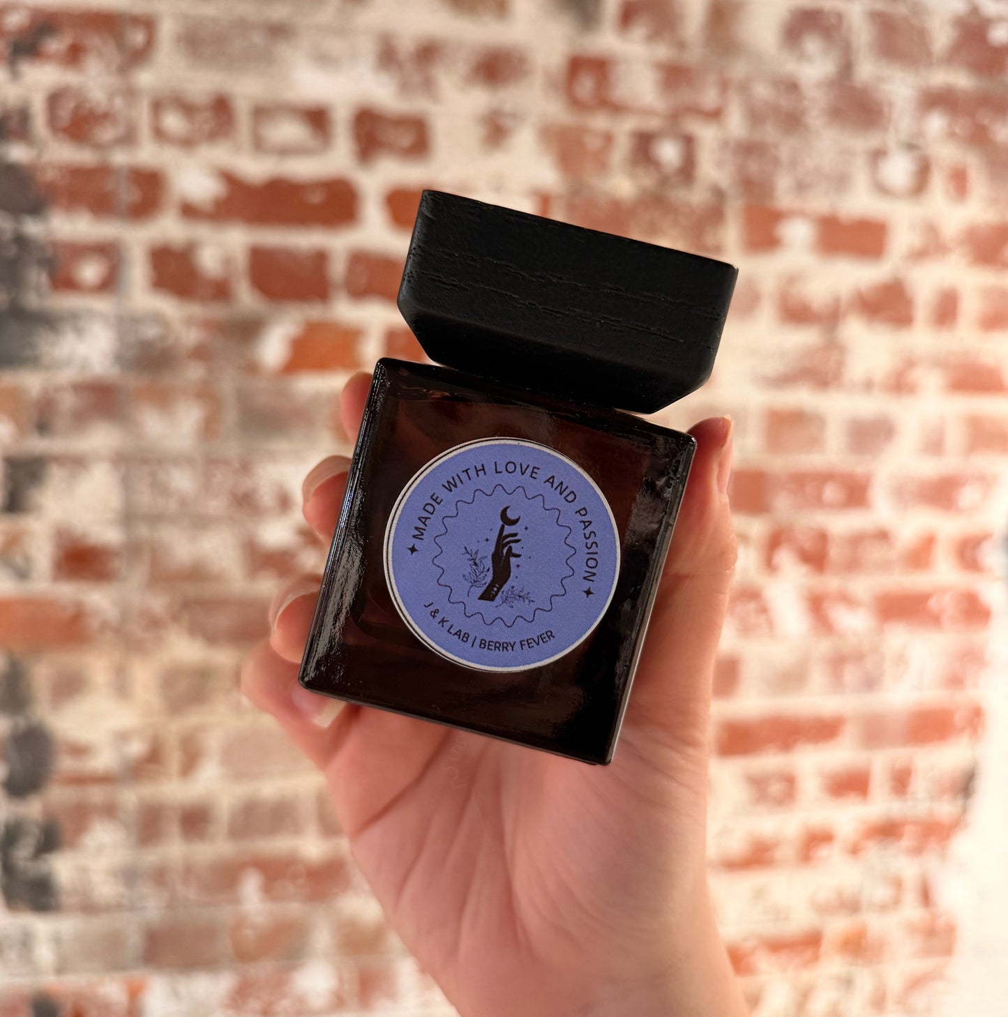 Reed diffuser in a dark glassware with a square wooden lid and purple round label. 