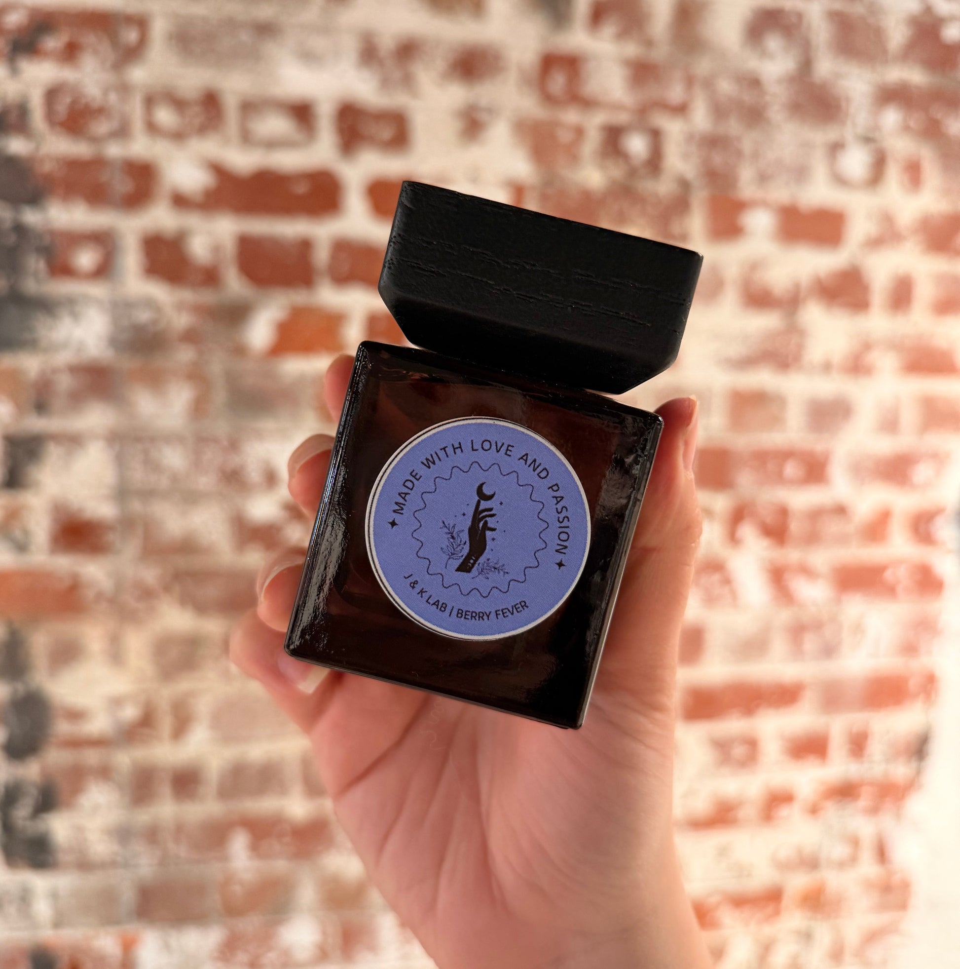 Reed diffuser in a dark glassware with a square wooden lid and purple round label. 