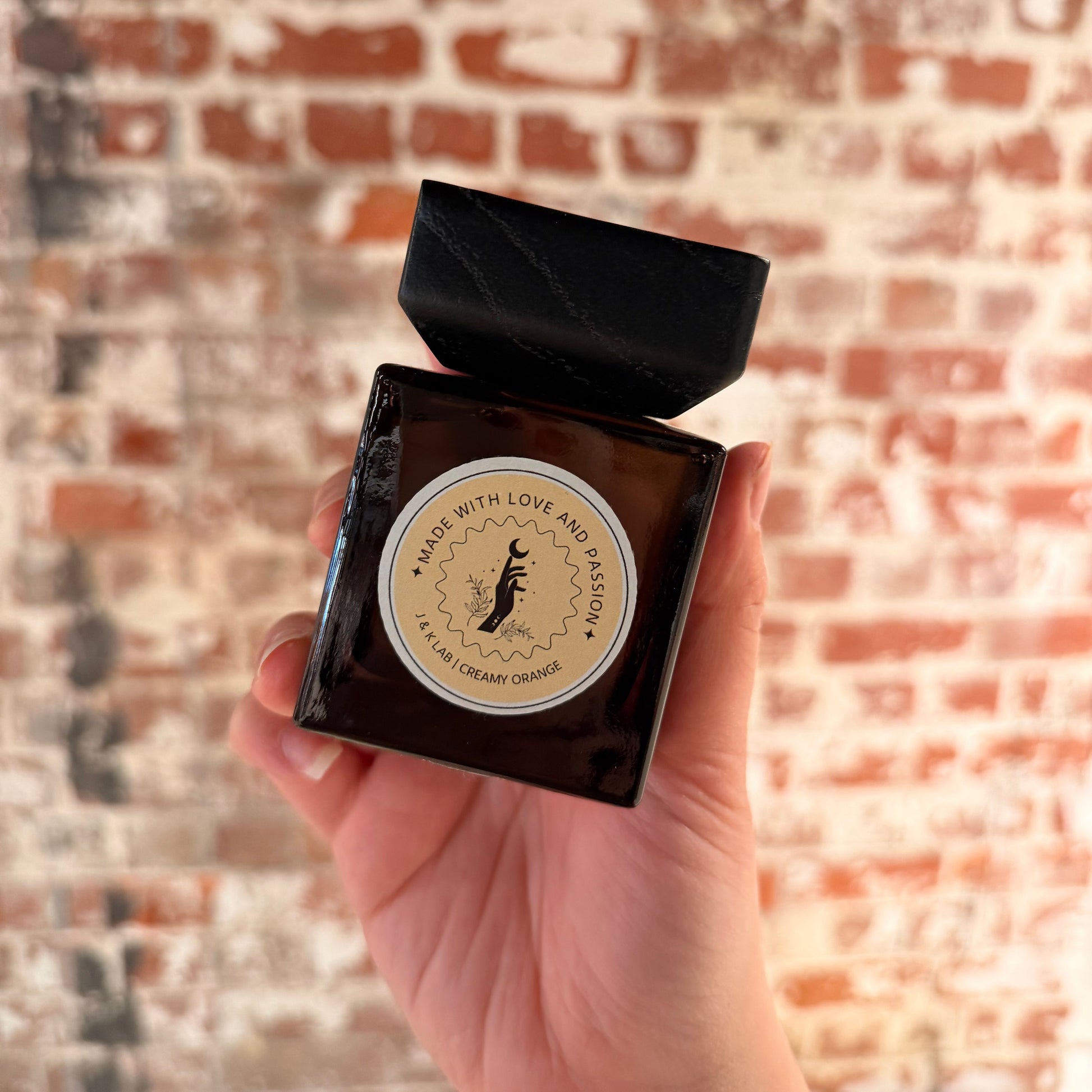 Reed diffuser in a dark glassware with a square wooden lid and light orange round label. 