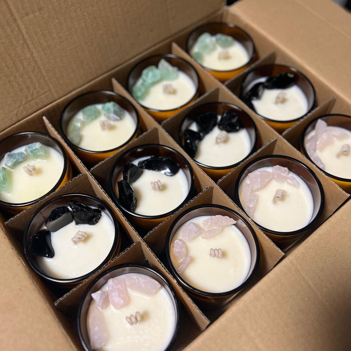 An open cardboard gift box containing multiple amber glass candles arranged in a grid. Each candle has creamy off-white wax and is topped with different polished gemstones in black, green, pink, and clear varieties.