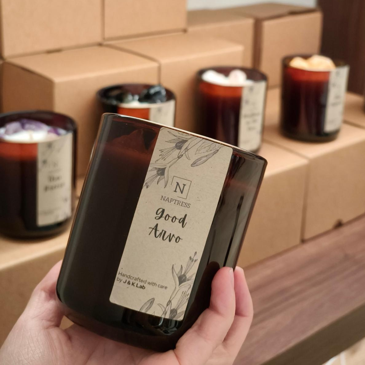 A hand holds a dark amber glass candle jar labeled 'Good Avro' by Napthess, with other candles and brown cardboard boxes blurred in the background.