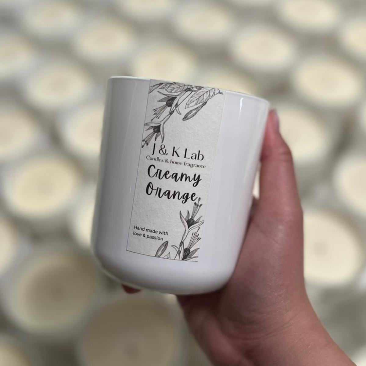 A hand holds a white candle jar labeled 'J & K Lab Creamy Orange' in front of a blurred background of many identical candles.