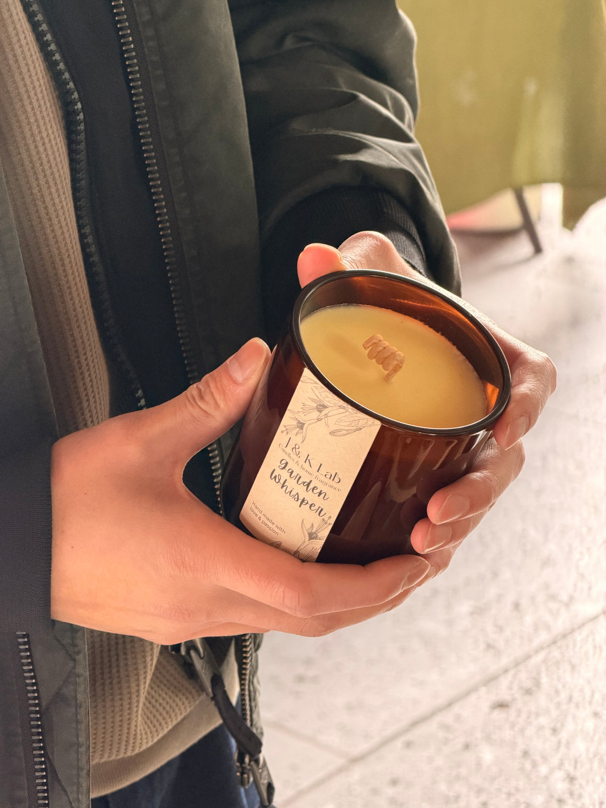 A person holding a brown amber glass jar candle with a cream label that reads 'J&K Lab Garden Whisper .' The candle is shown in hands against a softly blurred outdoor background.