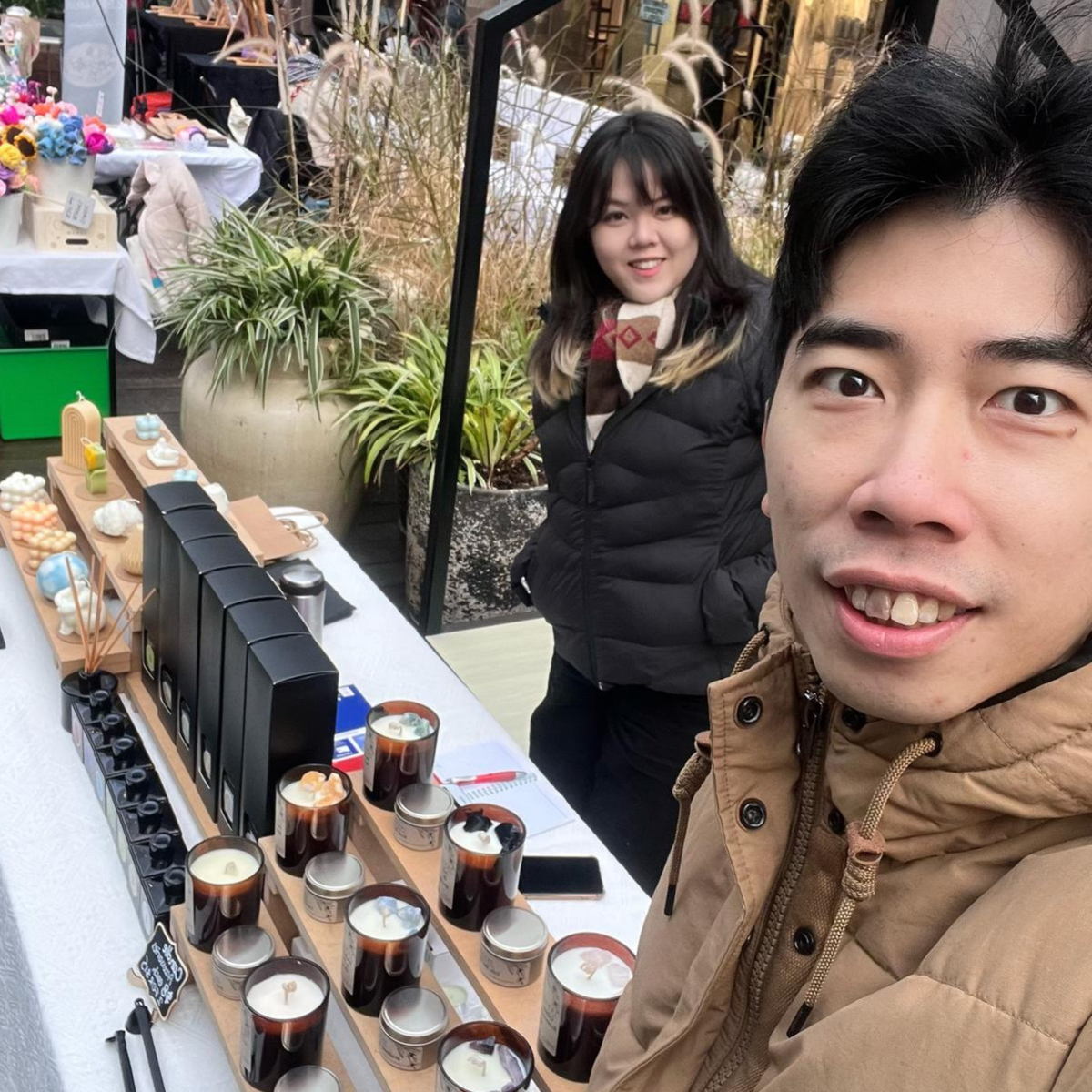 A man takes a selfie at an outdoor market booth displaying various scented candles, with a woman standing behind him.