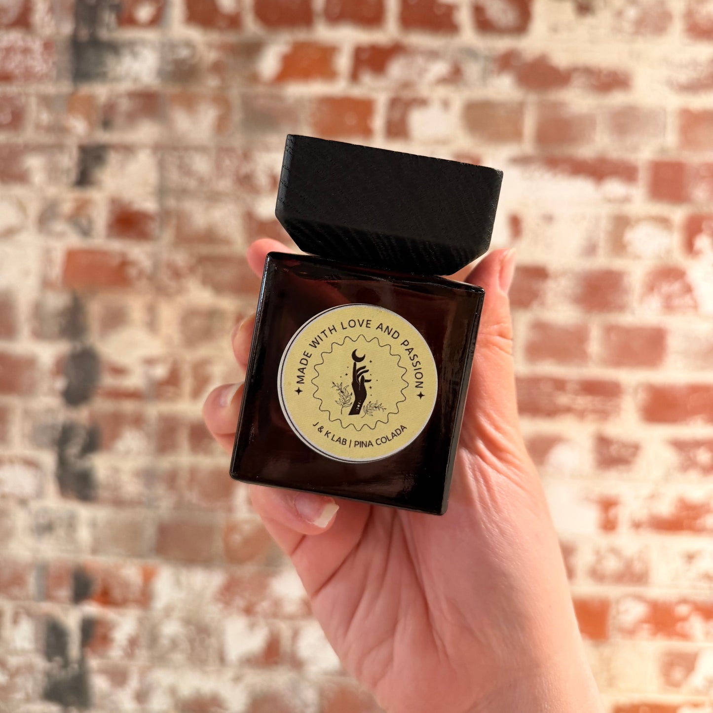 Reed diffuser in a dark glassware with a square wooden lid and baby yellow round label. 